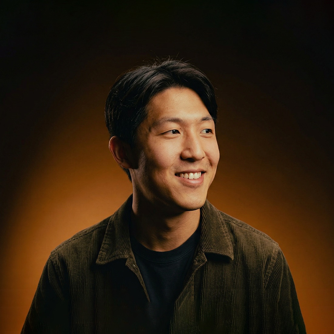 Alyosha Kim, smiling and looking off to the side, in a dark corduroy jacket against a warm amber backdrop.
