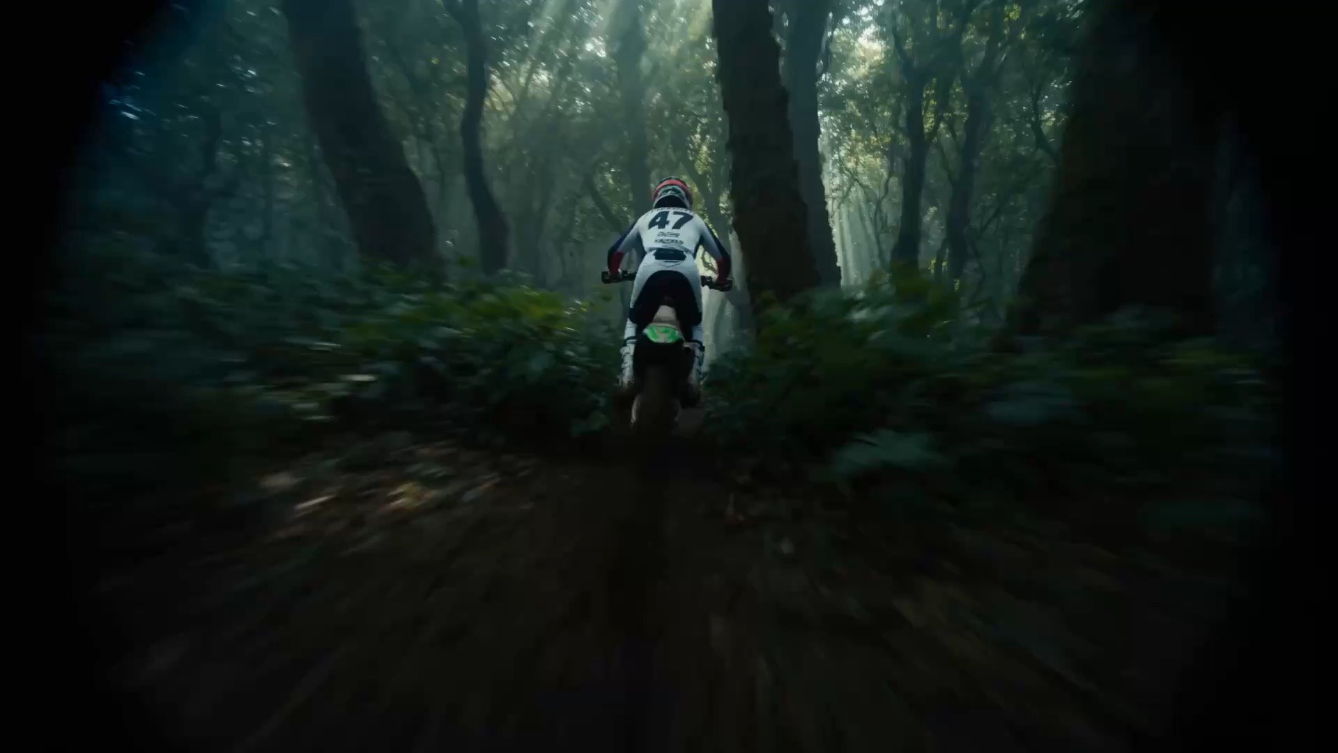 A dirt-bike rider in a white number 47 jersey races down a misty forest trail through shafts of light.