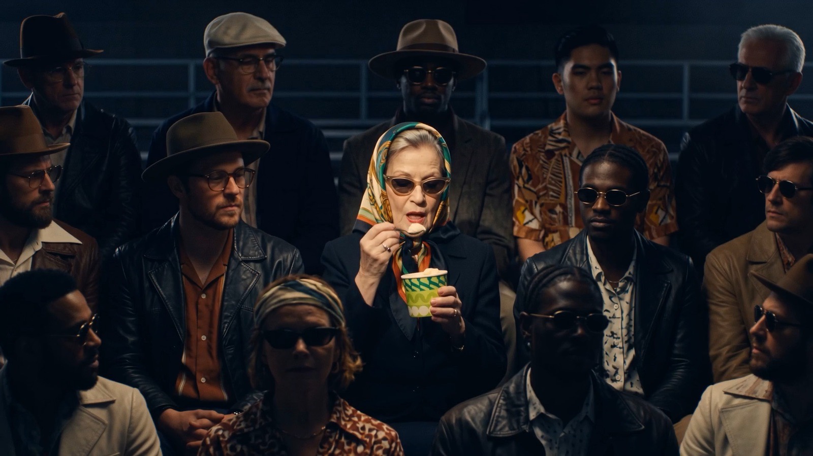 A dark grandstand of spectators in hats and sunglasses; one woman in a headscarf eats ice cream from a green tub.